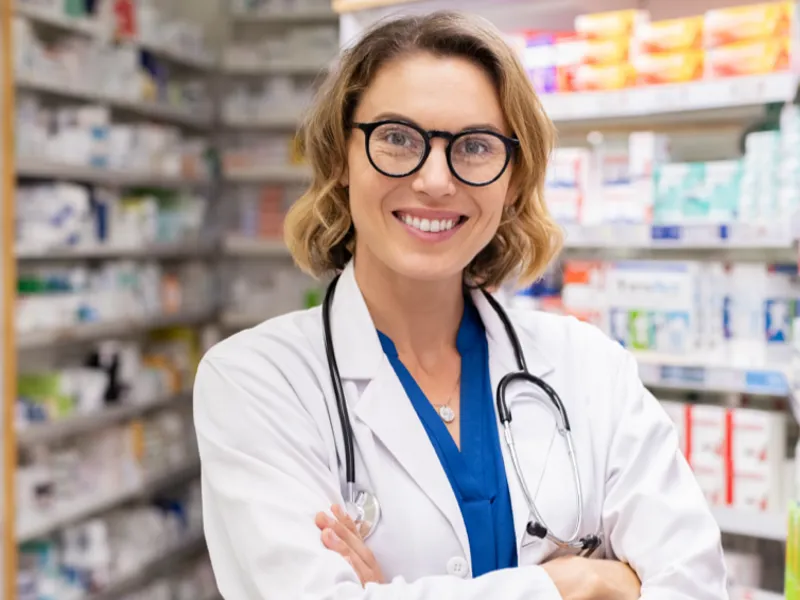 A smiling pharmacist standing in front of a wall full of medications.